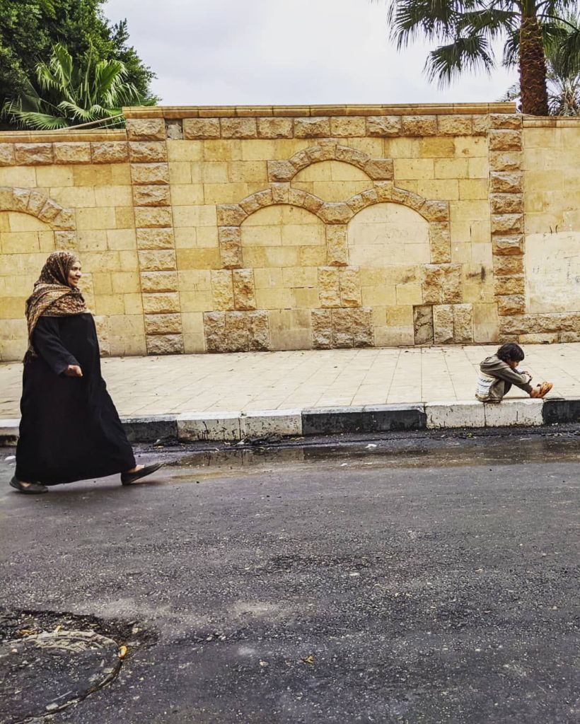 A woman walking down a street. A few feet ahead of her is a boy sitting on a sidewalk.