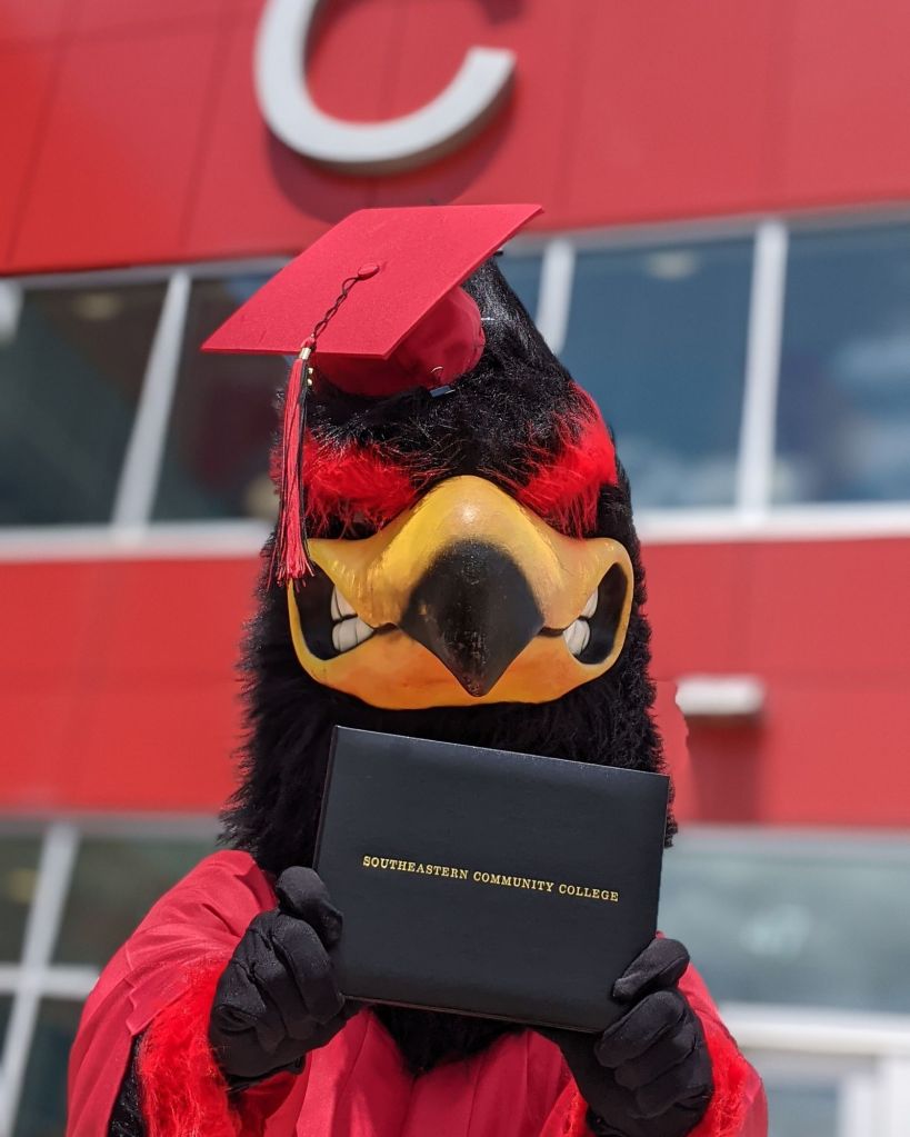 Bird mascot wearing a graduation outfit, holding a diploma.